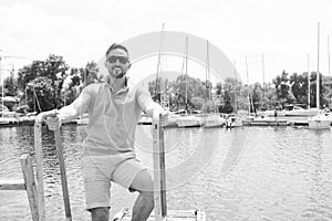 Man in polo and sunglasses laddering up on boat. Happy businessman on vacation with yachts on dock and wooden pier background
