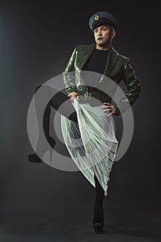 A man portraying a glamorous character in a dark green shiny tailcoat, cap, wedding veil and white petticoat, in heels