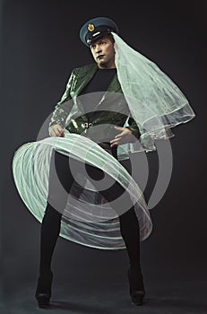 A man portraying a glamorous character in a dark green shiny tailcoat, cap, wedding veil and white petticoat, in heels