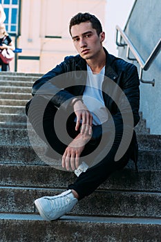man posing sits on the steps near railing. Handsome young man in stylish black clothes and white shoes posing near a railing