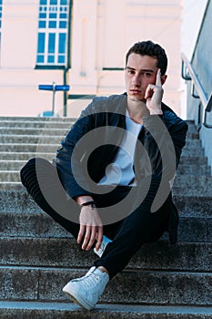 man posing sits on the steps near railing. Handsome young man in stylish black clothes and white shoes posing near a railing