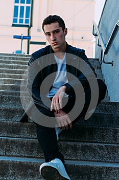 man posing sits on the steps near railing. Handsome young man in stylish black clothes and white shoes posing near a railing