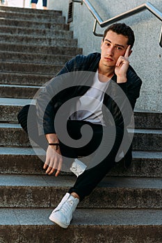 man posing sits on the steps near railing. Handsome young man in stylish black clothes and white shoes posing near a railing