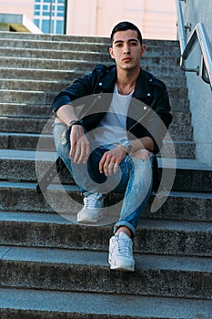 man posing sits on the steps near railing. Handsome young man in stylish black clothes and white shoes watch on hand