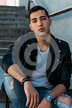 man posing sits on the steps near railing. Handsome young man in stylish black clothes and white shoes watch on hand