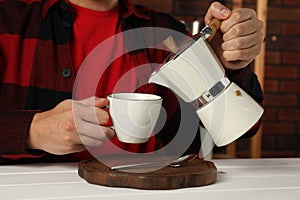 Man pouring aromatic coffee from moka pot into cup at white wooden table indoors, closeup