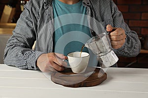 Man pouring aromatic coffee from moka pot into cup at white wooden table indoors, closeup