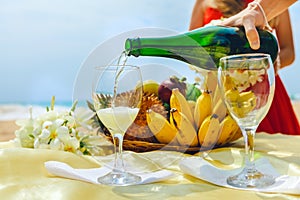 Man is pouring champagne into a glass on a background of a plate with fruit. Celebration on the beach