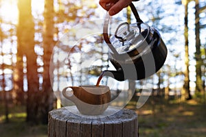 Man pouring coffee from kettle into a cup in the forest. Fresh coffee at campsite by the lake.