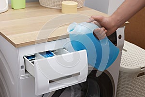 Man pouring fabric softener from bottle into washing machine indoors, closeup