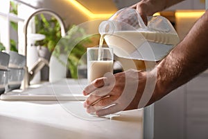 Man pouring milk from gallon bottle into glass at white countertop in kitchen, closeup