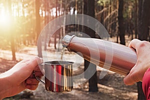 Man pouring tea from thermos in a metal cup while camping in pine forest.