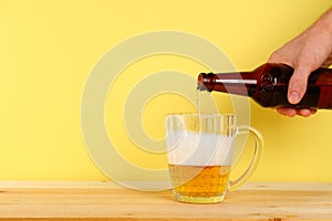 A man pours beer into a mug from a bottle on a yellow background on a wooden table. Copy space.