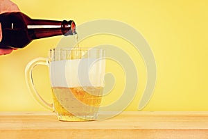 A man pours beer into a mug from a bottle on a yellow background on a wooden table. Copy space.