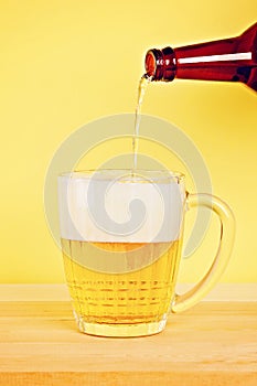 A man pours beer into a mug from a bottle on a yellow background on a wooden table. Copy space.