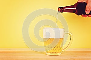 A man pours beer into a mug from a bottle on a yellow background on a wooden table. Copy space.