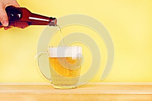 A man pours beer into a mug from a bottle on a yellow background on a wooden table. Copy space.