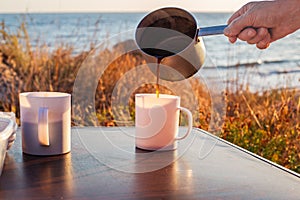 man pours freshly brewed coffee from an aluminum ladle into a mug on a tourist table, on the seashore. Travel and