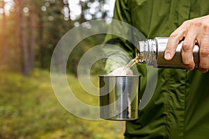 man pours hot drink from thermos flask into a metal cup in forest. nature tourism and camping gear concept