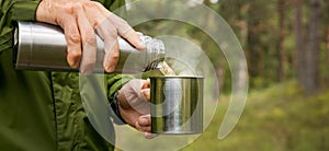 man pours hot tea from thermos flask into a mug in forest. nature tourism and camping concept. copy space