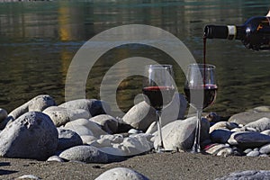 Man pours red wine from bottle into two glasses outdoors on rocky beach of mountain river