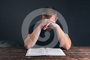 A man prays with folded hands. An open Holy Bible on the table. A young man with a beard.