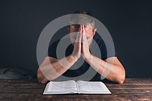 A man prays with folded hands. An open Holy Bible on the table. A young man with a beard.