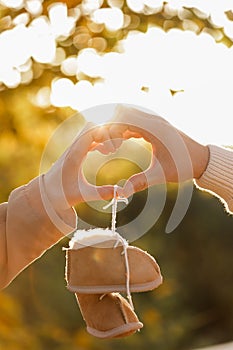 Man and pregnant wife are doing heart gesture with hands and holding warm baby shoes on nature autumn background