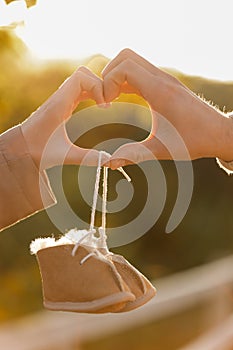 Man and pregnant wife are doing heart gesture with hands and holding warm baby shoes on nature autumn background