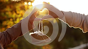 Man and pregnant wife are doing heart gesture with hands and holding warm baby shoes on nature autumn background