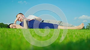 A man with a pregnant wife is lying on the green grass. Against the background of the blue sky