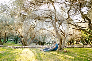 Man with a pregnant woman in a long blue dress lie on the grass under a sprawling olive tree in the park