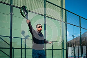 A man prepares for a backhand shot in padel tennis, with a focused expression and the court's fence shadow patterned