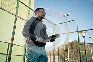 A man prepares for a backhand shot in padel tennis, with a focused expression and the court's fence shadow patterned