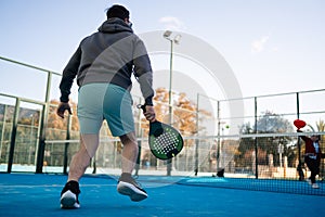 A man prepares for a backhand shot in padel tennis, with a focused expression and the court's fence shadow patterned
