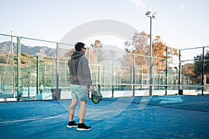 A man prepares for a backhand shot in padel tennis, with a focused expression and the court's fence shadow patterned