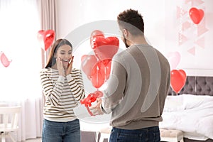 Man presenting gift to his girlfriend in bedroom decorated with heart shaped balloons. Valentine`s day celebration