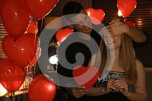 Man presenting gift to his girlfriend in room decorated with heart shaped balloons. Valentine`s day celebration