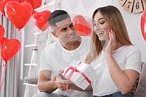 Man presenting gift to his girlfriend in room decorated with heart shaped balloons. Valentine`s day celebration