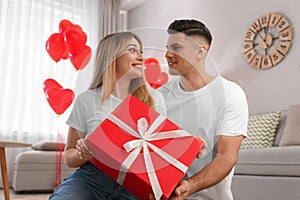 Man presenting gift to his girlfriend in room decorated with heart shaped balloons. Valentine`s day celebration