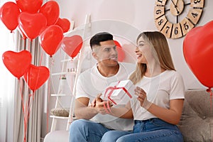 Man presenting gift to his girlfriend in room decorated with heart shaped balloons. Valentine`s day celebration