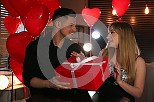 Man presenting gift to his girlfriend in room with heart shaped balloons. Valentine`s day celebration