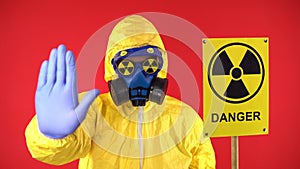 A man in a protective chemical suit, in a protective mask with a poster. Radiation shows a stop signal with his hand