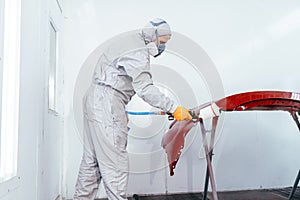 Man with protective clothes and mask painting car using spray compressor, red front bumper in chamber workshop.