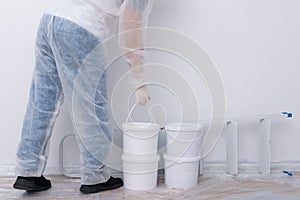 A man in protective clothing, bringing buckets of paint and a set of tools, against a white wall