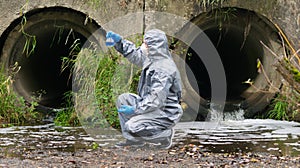 A man in protective clothing, looking at a tube of blue liquid, against the drainpipes