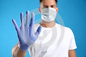 Man in protective face mask and medical gloves showing stop gesture against blue background, focus on hand