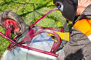 A man in a protective helmet welds a metal cart with an electrode. Agricultural machinery repair