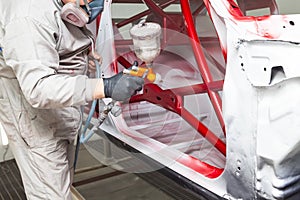 A man in protective overalls and a mask holds a spray bottle in his hand and sprays red paint onto the frame of the car body after