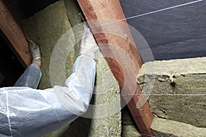 A man in a protective overalls puts mineral wool between the beam of the roof of the house for his warming from the cold